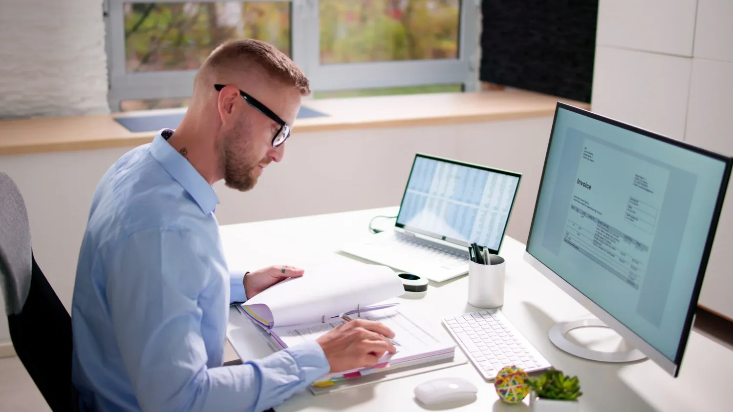 Ein junger Mann mit Brille sitzt an einem Schreibtisch mit einem PC und einem Laptop vor sich und schaut runter auf ein geöffnetes Buch.