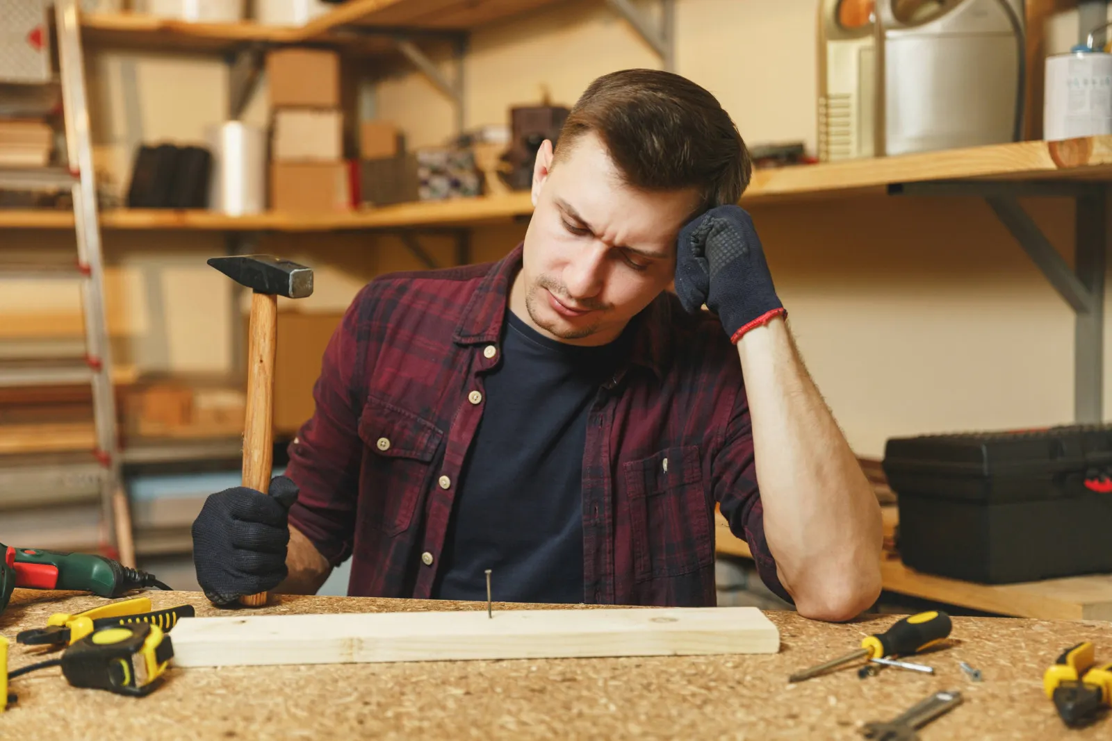 Ein Handwerker steht an einem Tisch mit einem Hammer in der einen Hand vor einem Holzbrett und stützt sich mit der anderen Hand ab. Er wirkt lustlos und unmotiviert seine Arbeit zu machen.