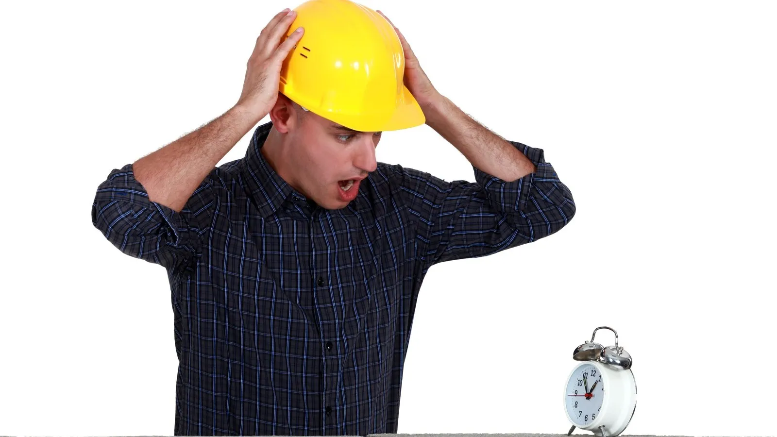Ein Handwerker mit gelbem Helm hält sich empört am Kopf und blickt auf einen weißen Wecker, der auf einer Mauer steht.