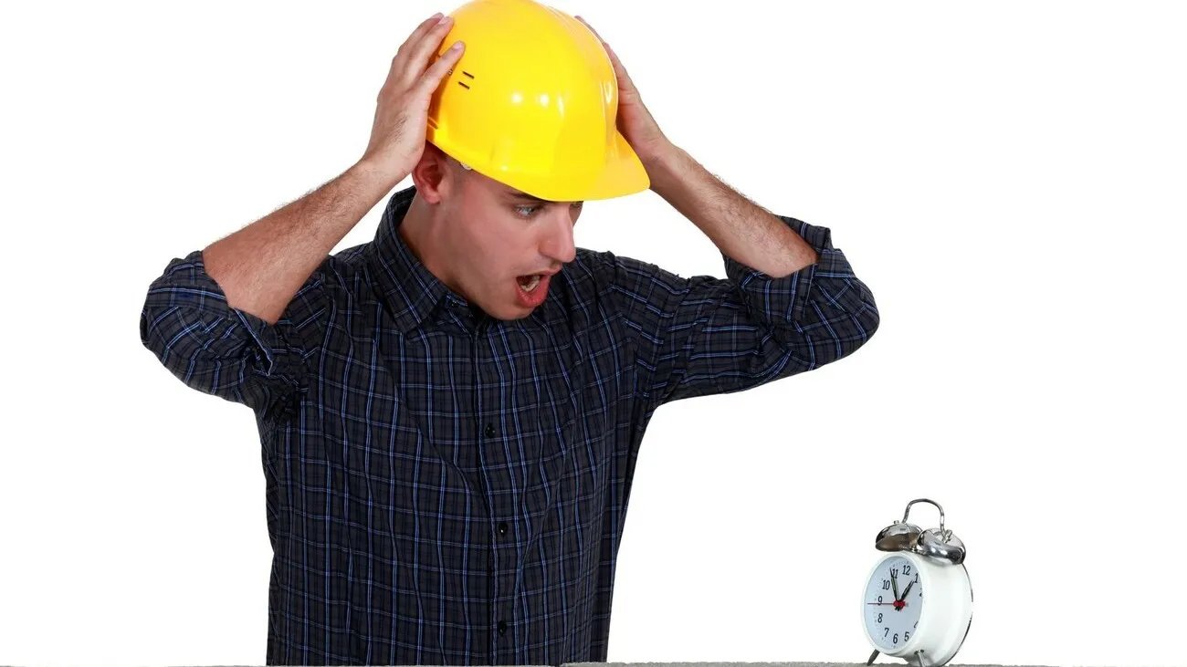 Ein Handwerker mit gelbem Helm hält sich empört am Kopf und blickt auf einen weißen Wecker, der auf einer Mauer steht.