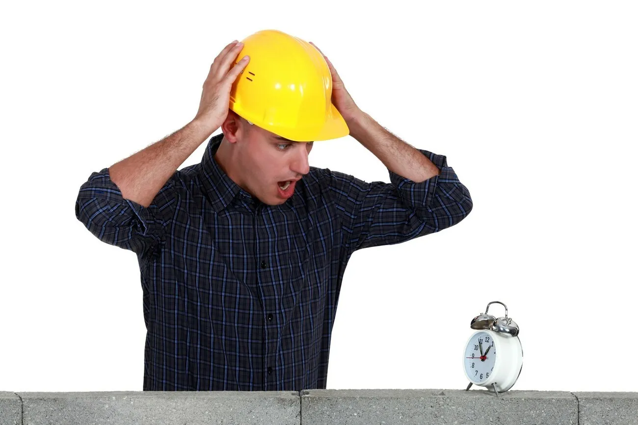 Ein Handwerker mit gelbem Helm hält sich empört am Kopf und blickt auf einen weißen Wecker, der auf einer Mauer steht.