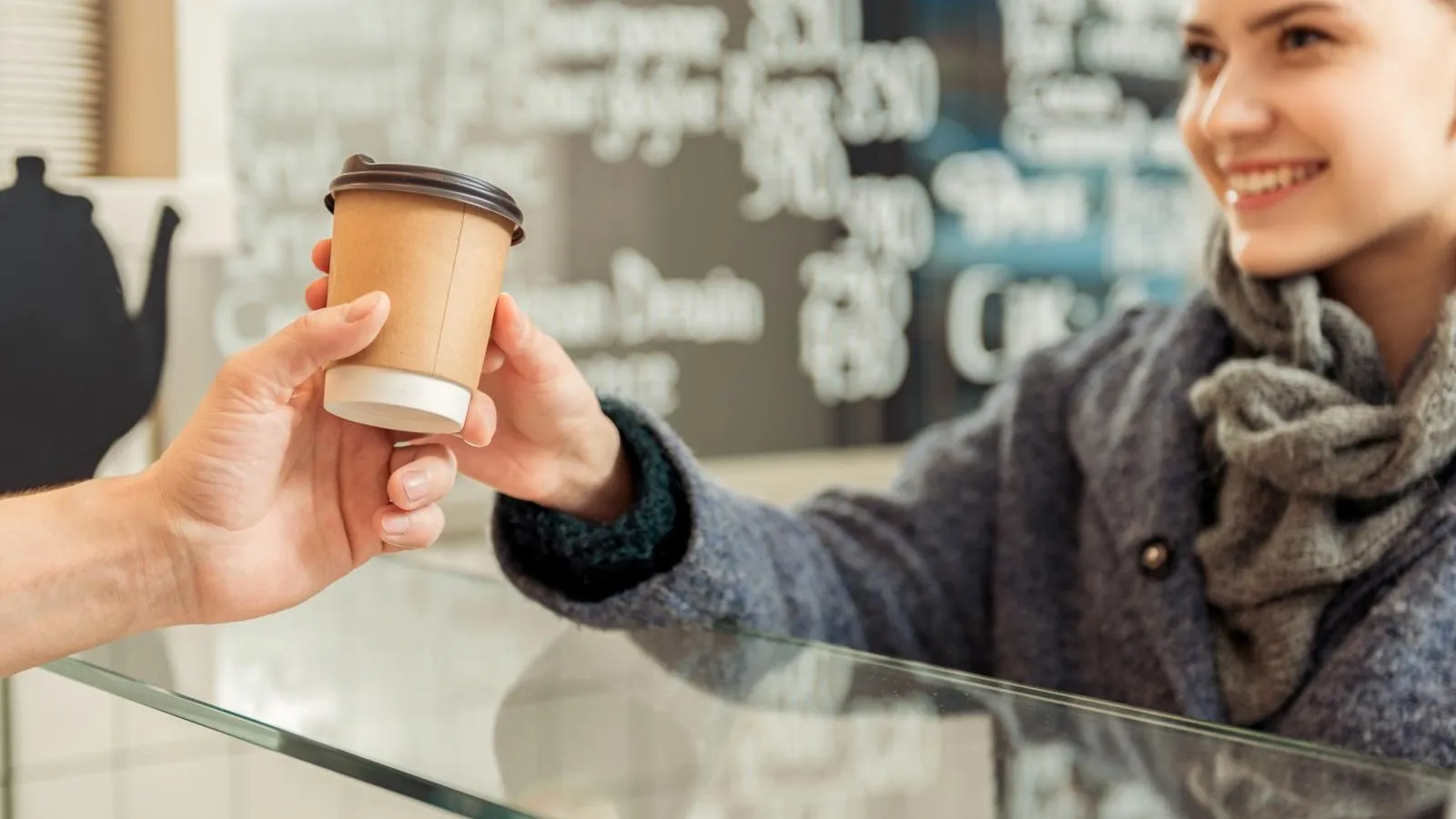 Ein Bäcker reicht einen Coffee To-Go über den Tresen. Ein Kundin nimmt den Becher per Hand entgegen.
