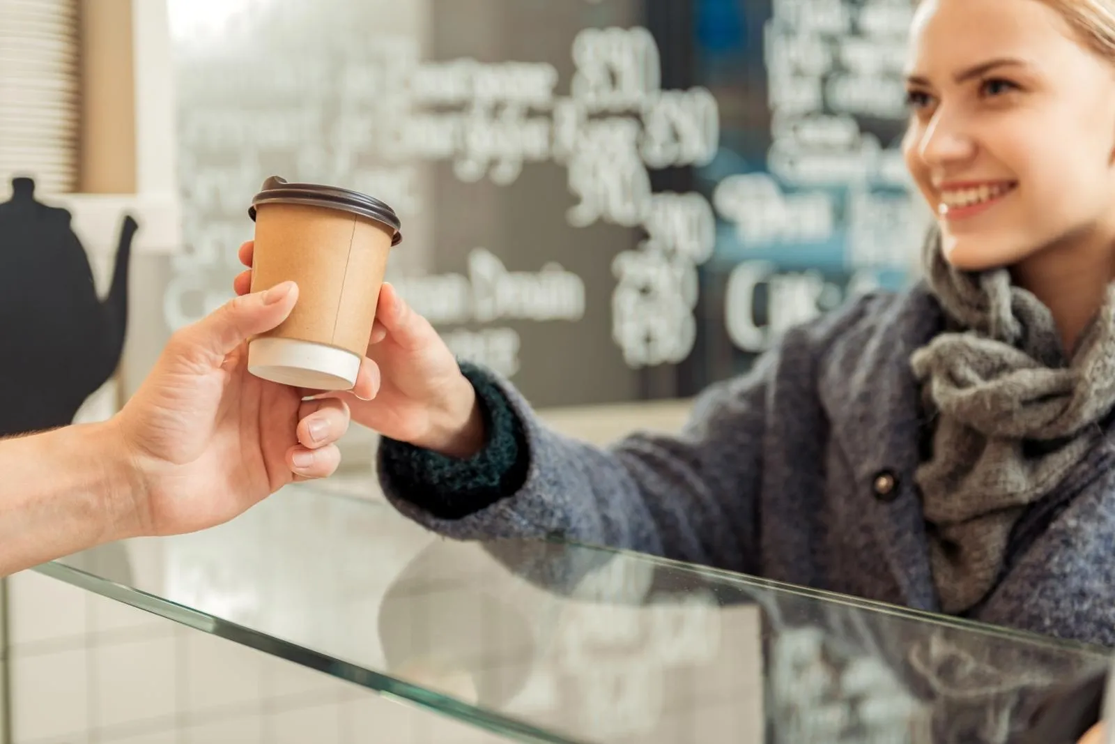 Ein Bäcker reicht einen Coffee To-Go über den Tresen. Ein Kundin nimmt den Becher per Hand entgegen.