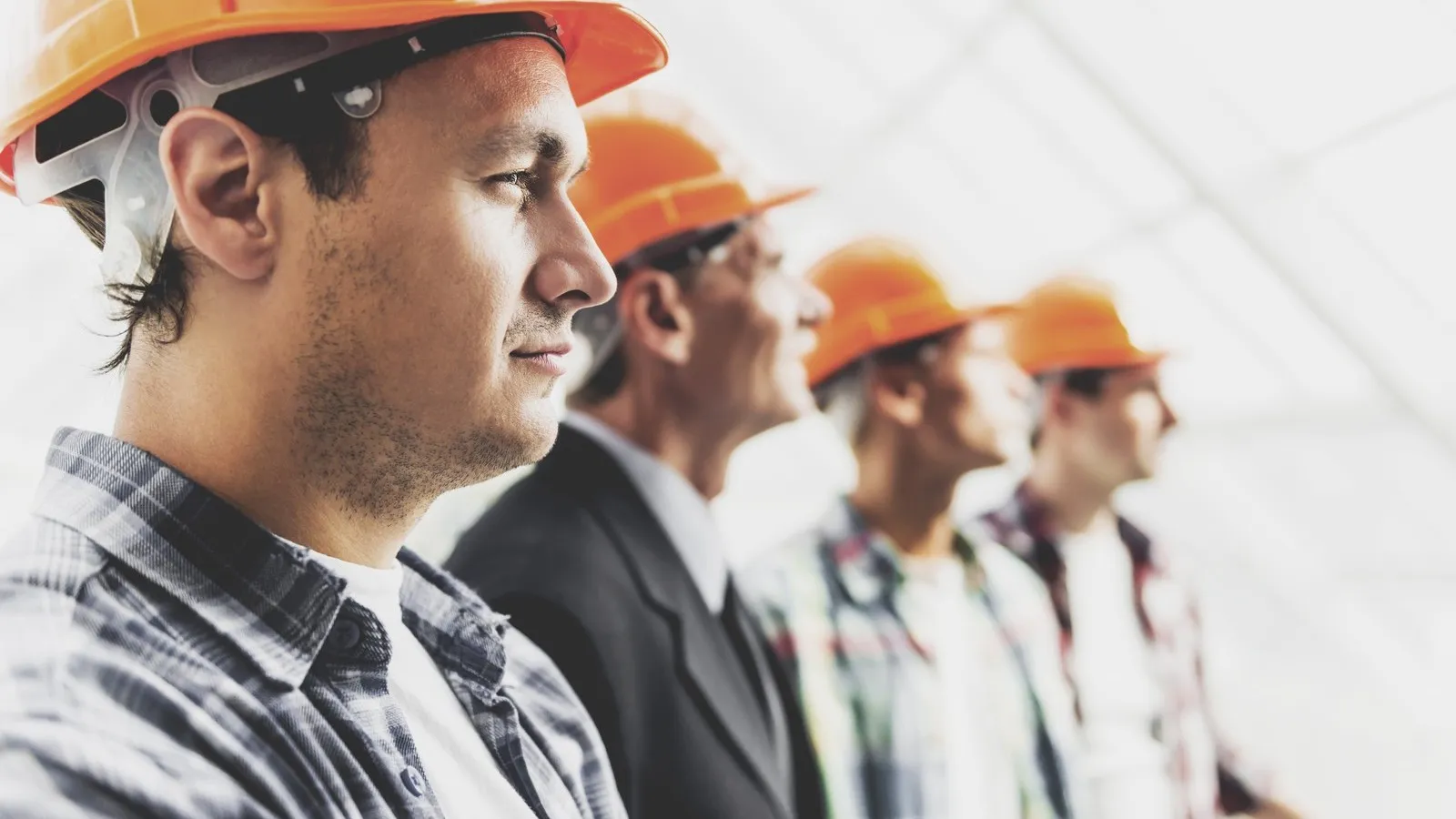 Bauarbeiter mit orangenen Schutzhelmen stehen in einer Reihe nebeneinander. Im Bildfokus steht ein Bauarbeiter von der Seite, die anderen sind dahinter weniger deutlich zu erkennen.