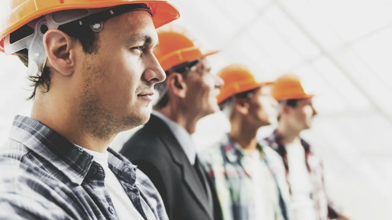 Bauarbeiter mit orangenen Schutzhelmen stehen in einer Reihe nebeneinander. Im Bildfokus steht ein Bauarbeiter von der Seite, die anderen sind dahinter weniger deutlich zu erkennen.