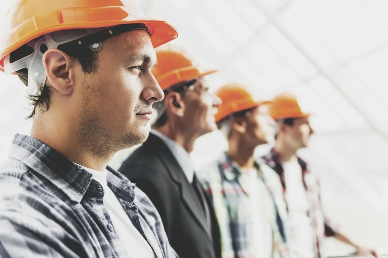 Bauarbeiter mit orangenen Schutzhelmen stehen in einer Reihe nebeneinander. Im Bildfokus steht ein Bauarbeiter von der Seite, die anderen sind dahinter weniger deutlich zu erkennen.