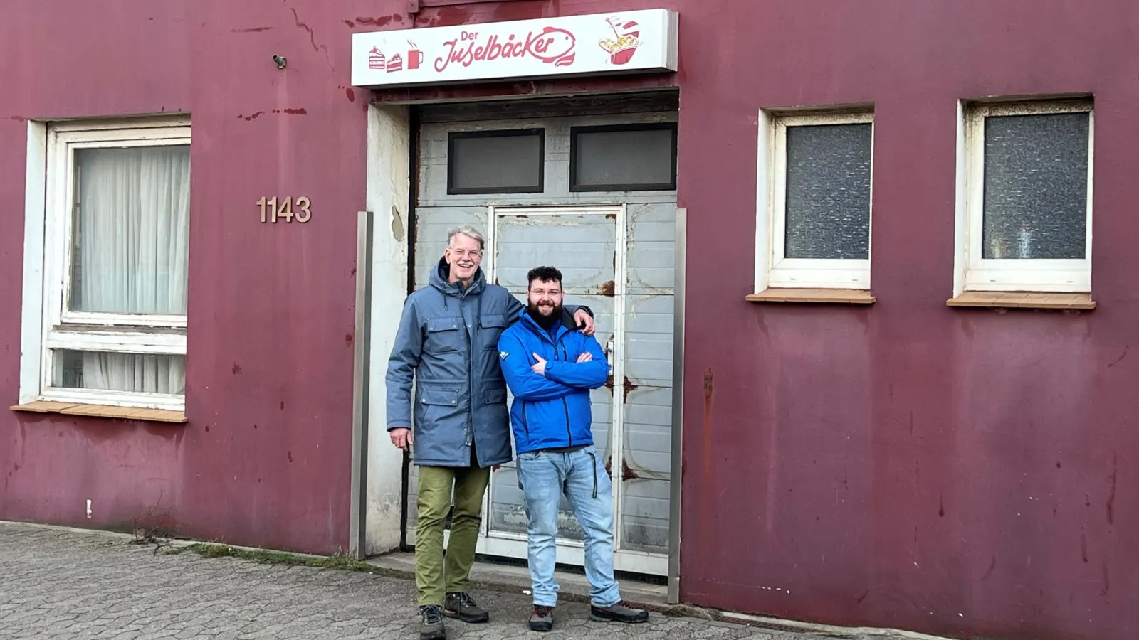 Bäckermeister Mario Peterscheck (links) und Adrian Wolf (rechts) stehen vor der Tür der alten Bäckerei in Helgoland. Die Wände des Gebäudes sind rötlich.