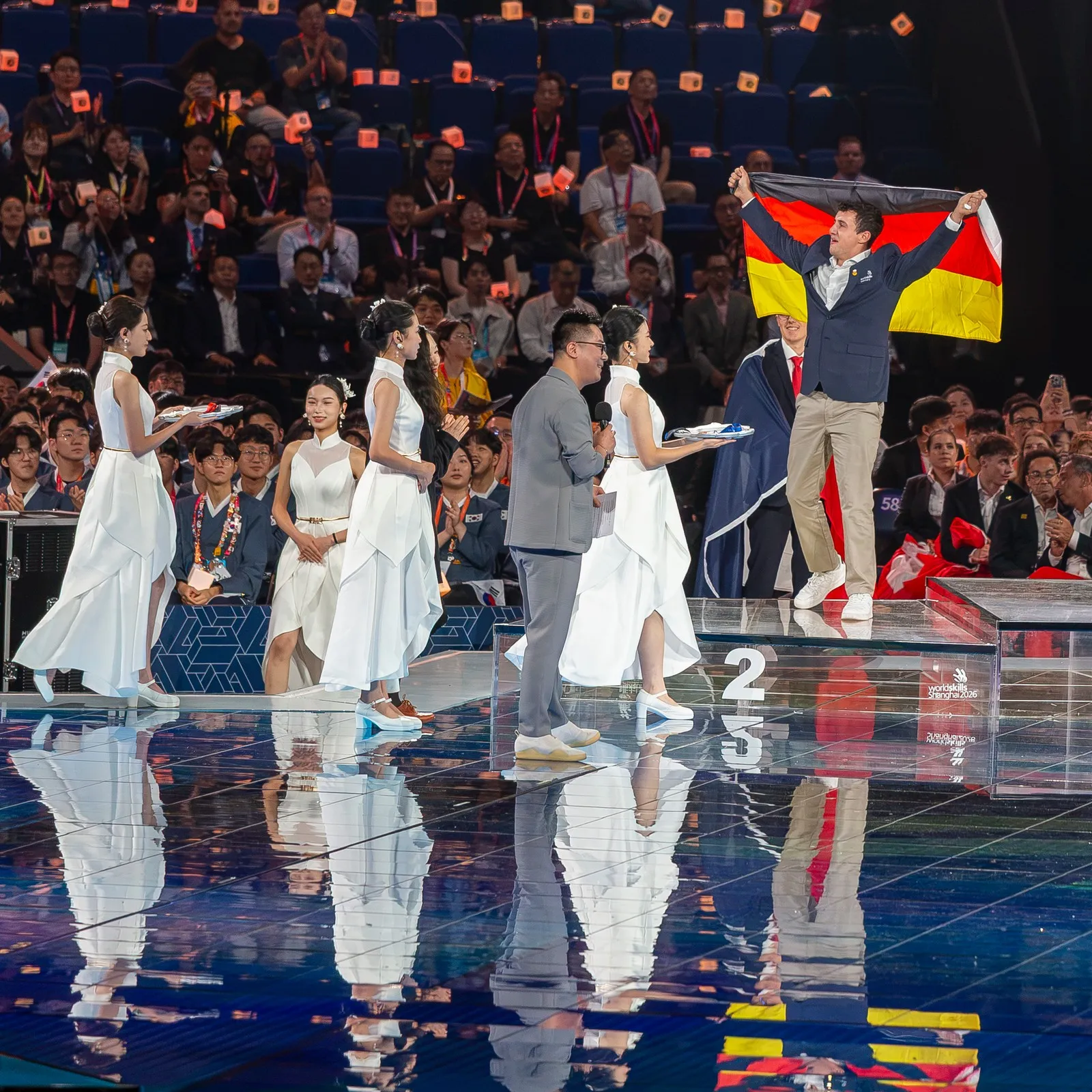 vier in weiß gekleidete Damen schreiten in Shanghai über eine Bühne, um den Siegern der WorldSkills die Medaillen zu überreichen. In den Händen halten sie jeweils ein Tablett, auf dem die Medaillen bereit liegen. Auf dem Podest hält Florian Dorer die Deutschlandfahne in die Luft. Er steht auf dem Treppchen des zweiten Platzes.