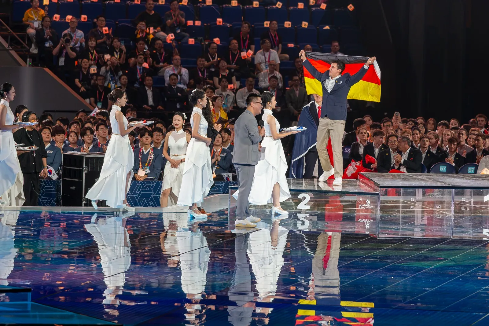 vier in weiß gekleidete Damen schreiten in Shanghai über eine Bühne, um den Siegern der WorldSkills die Medaillen zu überreichen. In den Händen halten sie jeweils ein Tablett, auf dem die Medaillen bereit liegen. Auf dem Podest hält Florian Dorer die Deutschlandfahne in die Luft. Er steht auf dem Treppchen des zweiten Platzes.
