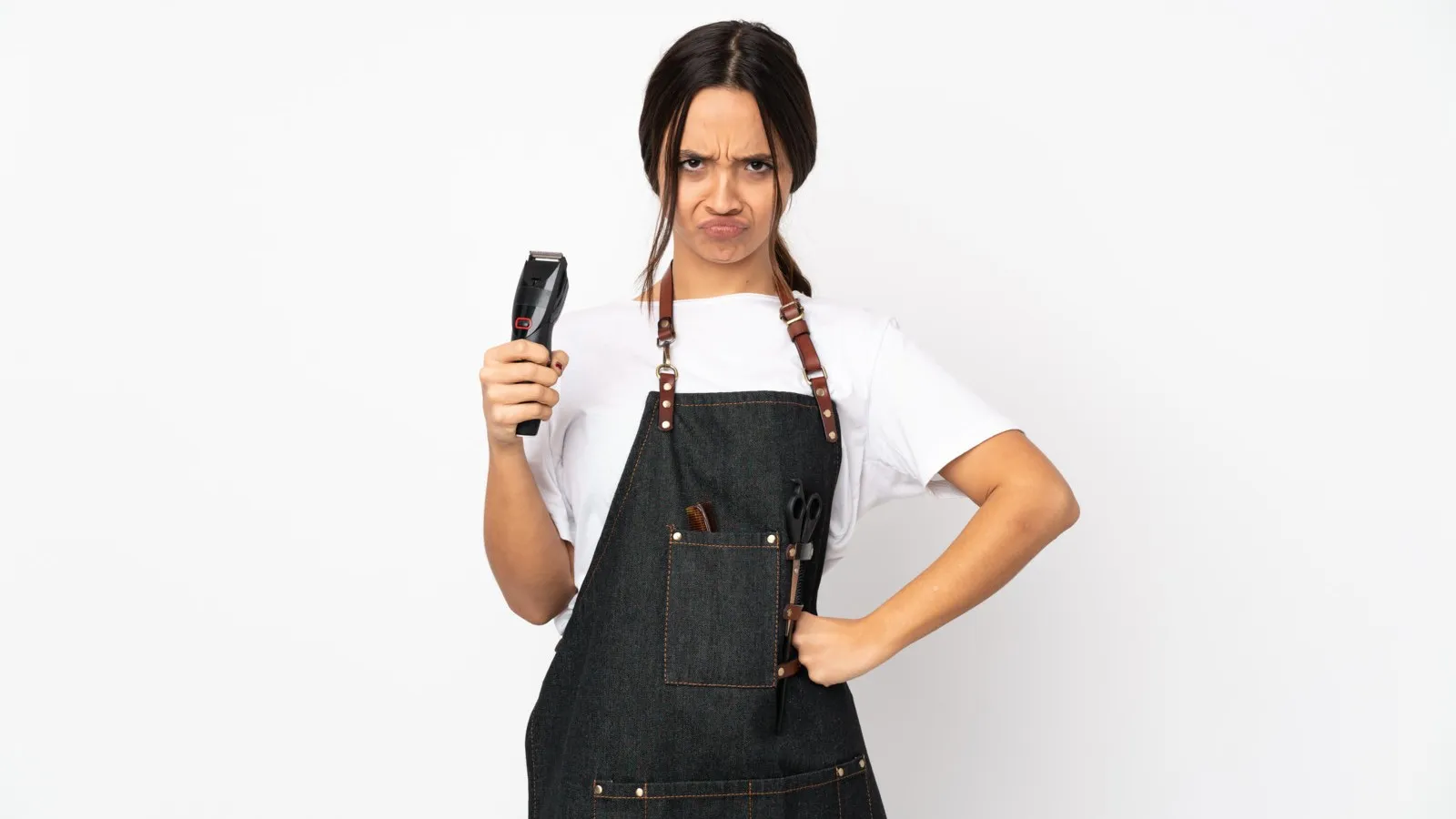 Eine wütende Friseurin hält einen Trimmer in der rechten Hand hoch und stützt sich mit ihrer linken Hand an der Hüfte ab.