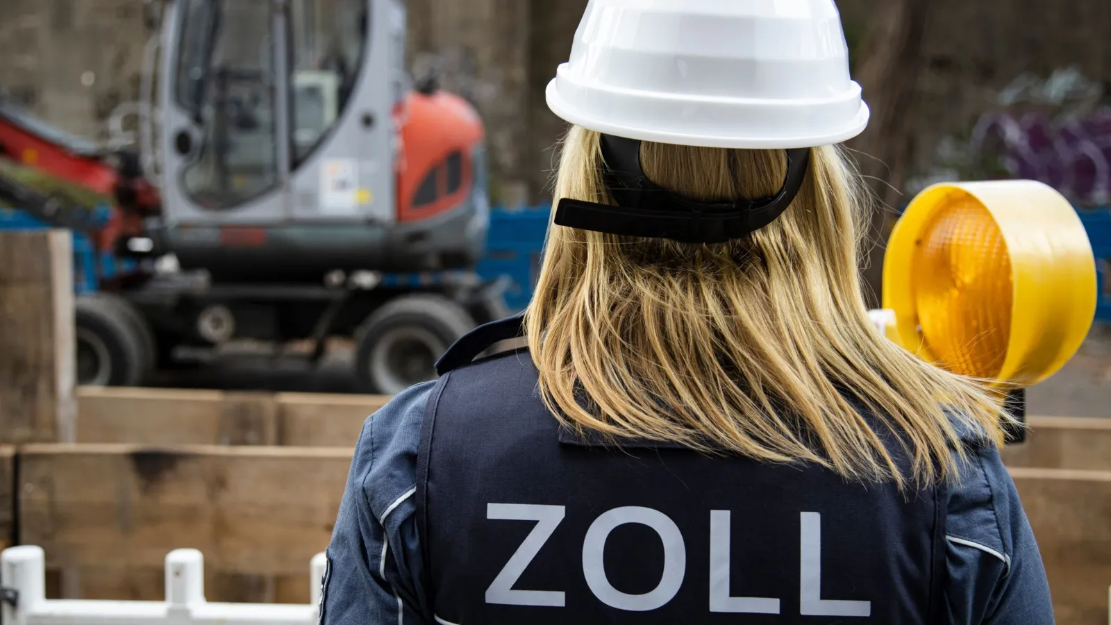 Eine Frau mit einem weißen Schutzhelm und einer Weste mit der Aufschrift "Zoll" ist von hinten zu sehen. Im Hintergrund ist eine Baustelle.